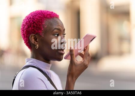 Schwarze Frau mit kurzgefärbtem Haar, die Sprachnachrichten mit einem Smartphone aufnimmt, das tagsüber auf der Straße steht Stockfoto