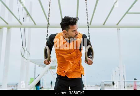 Hispanischer Mann, der Gymnastikringe in einem Fitnessraum in der Nähe des Strandes ausführt und während eines Workouts wegblickt. Dieses Fitness-Bild zeigt Krafttraining, Outdoor-Übungen und einen aktiven Lebensstil. Stockfoto
