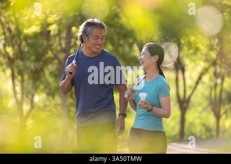 Zwei lächelnde chinesische Paare mittleren Alters laufen zusammen in einem sonnigen Park und halten Badmintonschläger. Die Szene fängt einen fröhlichen Moment der Outdoor-Fitness und Zweisamkeit in einer natürlichen grünen Umgebung ein. Ideal für Lifestyle-Bilder mit aktivem Altern, Freizeitaktivitäten und gesundem Leben. Stockfoto