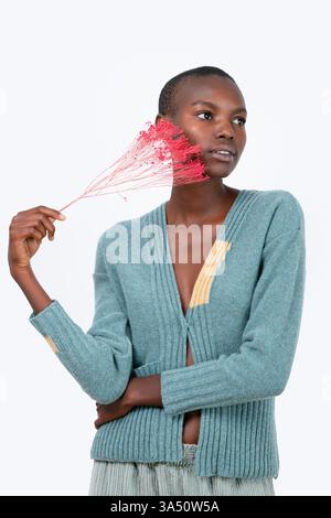 Nachdenkliche junge afroamerikanische Frau in einem blauen Pullover hält ein künstliches rosafarbenes Pflanzendekor und blickt vor einem weißen Hintergrund weg. Dieses Studio-Lifestyle-Bild passt zu Wohnkultur und modernem Innendesign. Perfekt für Design-, Wellness- und Wohnzimmerdekor-Kampagnen. Stockfoto