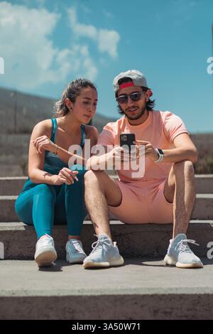 Der hispanische Mann zeigt seiner Freundin mit gemischter Rasse ein Smartphone, während er an einem sonnigen Tag auf einer Parktreppe sitzt. Dies fördert den Lebensstil von Paaren, mobile Technologie und Freizeit im Freien mit warmem, natürlichem Licht. Stockfoto