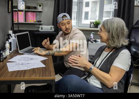 Ein asiatischer Tätowierer mit Mütze sitzt mit einer Kaukasierin mittleren Alters an einem Tisch und bespricht vor der Sitzung eine Tätowierungsskizze. In einem Studio fängt der Moment Kreativität, Konversation und professionelle Tattoo-Kunst ein. Ideal für Körperkunst, Tätowierstudios und die Zusammenarbeit zwischen Kunden und Künstlern. Stockfoto
