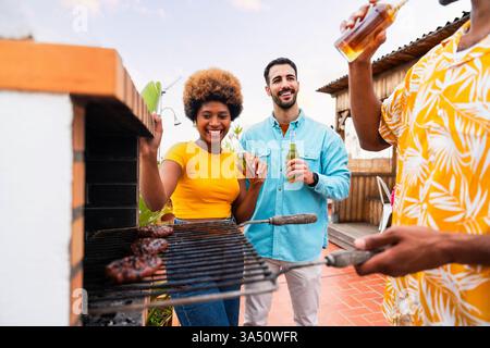 Fröhliche, vielfältige Gruppe von Freunden, die tagsüber einen Grill auf dem Dach genießen. Eine helle soziale Szene mit Speisen und Getränken, perfekt für Lifestyle- und Urban Living-Kampagnen. Stockfoto