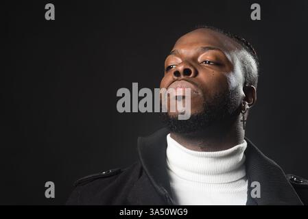 Serious Black man in einem weißen Rollkragenpullover und einem schwarzen Trenchcoat steht vor einem dunklen Studio-Hintergrund. Das Porträt vermittelt Selbstvertrauen und zeitgenössische Mode, die sich für urbane Lifestyle, Branding und Kampagnen im Executive-Stil eignet. Stockfoto