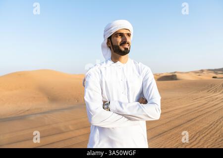 Ein erfolgreicher Mann aus dem Nahen Osten mit Kandura steht in den Wüstendünen. Das Bild zeigt ein traditionelles Outfit in einer beeindruckenden Wüstenlandschaft. Ideal für Unternehmensprofile, Führungsposition auf Golfmärkten und kulturelle Zusammenhänge im Nahen Osten. Stockfoto
