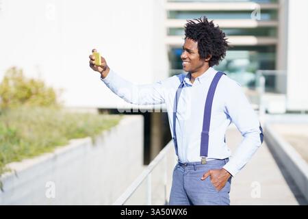 Fröhlicher afroamerikanischer Unternehmer mit einem hübschen Lächeln macht ein Selfie in der Nähe eines Bürogebäudes. Die urbane, geschäftsbereite Atmosphäre macht dieses Image perfekt für Unternehmensprofile, Startup-Funktionen oder Social-Media-Inhalte. Die Verwendung von Smartphones und der selbstbewusste Ausdruck vermitteln modernes Unternehmertum und beruflichen Erfolg. Stockfoto