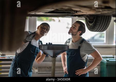 Schwarze und kaukasische Mechaniker untersuchen ein Auto unter einem angehobenen Fahrzeug in einer Autowerkstatt. Die Szene zeigt Teamwork, Vielfalt und professionellen Automobilservice. Ideal für Fahrzeugwartung, Werkstatt und Kfz-Servicekampagnen. Stockfoto