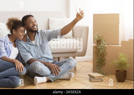Fröhliches schwarzes Paar posiert für ein Selfie in ihrer neuen Wohnung und feiert den Hausbesitz. Ideal für Immobilien, Hypotheken, bewegende Ankündigungen oder Lifestyle-Geschichten. Stockfoto