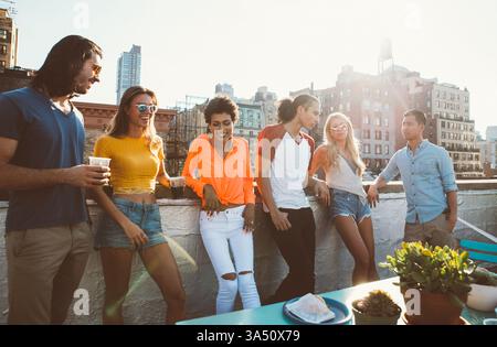 Verschiedene Gruppen von Freunden, die sich an einem sonnigen Tag auf einem Dachgeländer lehnen. Diese Outdoor-Aufnahme für den urbanen Lifestyle vermittelt Freundschaft, Feier und soziale Beziehungen. Stockfoto