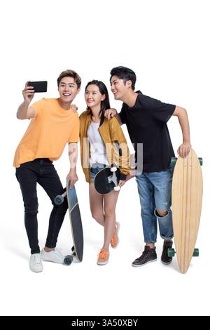Lächelnde chinesische Freunde machen ein Selfie auf einem Smartphone, während sie mit Skateboards im Studio stehen. Die Gruppe verströmt Freundschaft und Jugendkultur in einem hellen, lässigen Bild. Ideal für Lifestyle, Technologie und Social-Media-Inhalte mit Freunden und urbanem Spaß. Stockfoto
