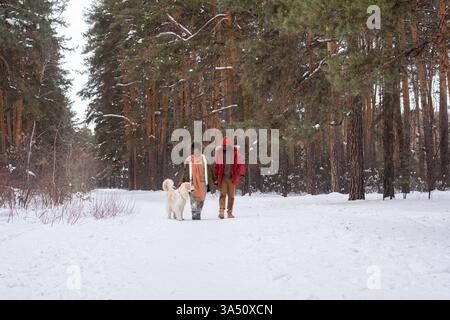Fröhliches afrikanisches Paar, das mit ihrem Hund durch einen Winterwald spaziert, an einem Wochenendausflug. Die Szene vermittelt Zweisamkeit, Outdoor-Aktivitäten und einen warmen, fröhlichen Lebensstil. Stockfoto