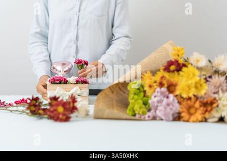 Die hispanische Blumenistin arrangiert rosa und weiße Nelken in einer Holzkiste, die mit einer Schleife in einem Blumengeschäft gebunden ist. Diese fokussierte Komposition unterstreicht das Handwerk und die Präsentation von Geschenken, perfekt für Blumendesign, Hochzeitsdekoration oder Ladenszene. Frische Blüten und warmes Licht sorgen für eine einladende, romantische Stimmung. Stockfoto