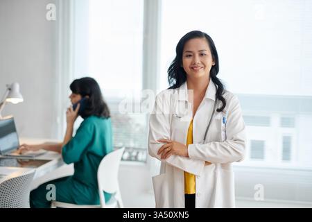 Lächelnde asiatische Ärztin in einem weißen Labormantel steht in einer klinischen Umgebung, mit einem Stethoskop zur Hand. Dieses selbstbewusste Porträt vermittelt Gesundheitskompetenz und mitfühlende Patientenversorgung für die Kommunikation in Krankenhaus und Klinik. Geeignet für medizinisches Marketing, Anbieter-bios oder Gesundheitskampagnen. Stockfoto