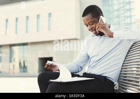 Ein professioneller afroamerikanischer Geschäftsmann in einem modernen Büro spricht mit einem Mobiltelefon, während er Dokumente überprüft. Ein Versicherungsvertreter prüft Papiere und berät auf einem Smartphone, wobei er den Führungsstil und die digitale Konnektivität in einem städtischen Geschäftsumfeld hervorhebt. Stockfoto