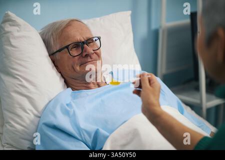Lächelnder älterer Mann, der auf dem Bett liegt und seiner Frau lächelt, während sie ihn füttert Stockfoto