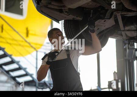 Ein afroamerikanischer Mechaniker zieht die Schrauben an einem angehobenen Fahrzeug in einer Autowerkstatt fest. Ein professioneller, praxisnaher Ort, der für Kfz-Dienstleistungen, Fahrzeugpflege oder Werkstattkampagnen geeignet ist. Stockfoto