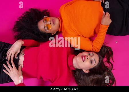 Hispanische und kaukasische Frauen liegen auf einem rosafarbenen Studioboden eng zusammen, um eine entspannte Freundschaftsszene zu erleben. Ideal für Lifestyle, Freundschaft und Porträtkonzepte im Studio. Stockfoto