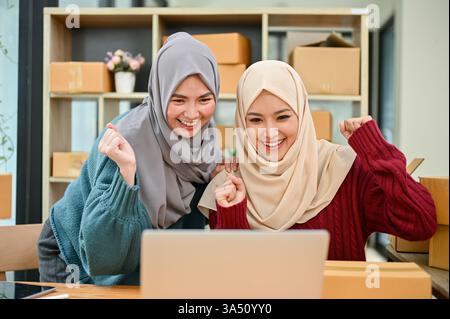 Ein junges muslimisches Team von Online-Unternehmen feiert gemeinsam, nachdem sie große Bestellungen auf ihrer Shopping-Website erhalten haben. Dieser vielfältige Startup-Moment zeigt Unternehmertum, E-Commerce und Frauenführung in einem muslimischen Kontext. Perfekt für Marketingkampagnen zum Online-Einzelhandel, zu technischen Startups und zum Erfolg kleiner Unternehmen. Stockfoto
