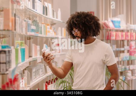Bärtiger Mann mit Afro liest Zutaten auf einem Arzneimittelflaschenetikett in einem Apothekengang. Dieses Bild im Gesundheitswesen passt zu medizinischen, pharmazeutischen und gesundheitlichen Kontexten. Stockfoto