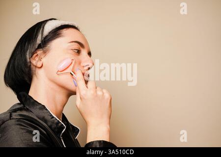 Schönheitsbehandlungen zu Hause. In einem Haarreif, der ihr Haar hält, gibt sich ein Mädchen eine Gesichtsmassage mit einem Gouachestein. Ein Gesichtsmassageroller in A Stockfoto