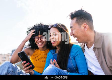 Fröhliche indische Frau sitzt mit Freunden aus Hispanic und Asien am sonnigen Strand und schaut auf ein Smartphone. Eine multikulturelle Reiseszene, in der Freundschaft, geselliges Teilen und Sommerspaß am Meer hervorgehoben werden. Ideal für Lifestyle, Reisen und multikulturelle Kampagnen. Stockfoto