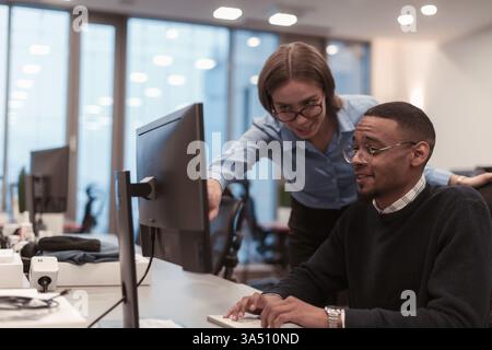 Lächelnder Schwarzer Mann, der am Schreibtisch sitzt und an seinem Computer arbeitet und mit einer kaukasischen Kollegin redet, die sich über die Schulter beugt und etwas auf die... Stockfoto