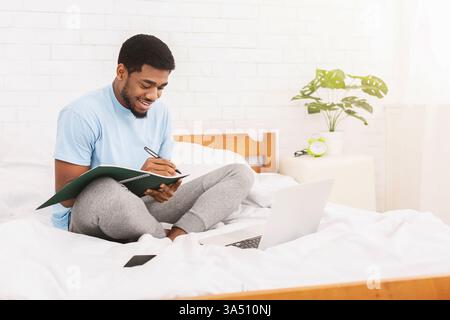 Millennial Black man studiert online im Bett, macht Notizen in einem Copybook, während er einen Laptop benutzt und schaut sich ein Webinar an. Dieses Foto ist ideal für E-Learning, Fernstudium und Bildmaterial zu Hause. Stockfoto