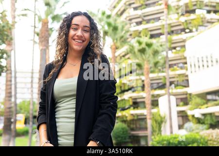 Die selbstbewusste junge hispanische Geschäftsfrau in einer stilvollen schwarzen Jacke blickt aus einem niedrigen Winkel auf die Kamera vor einer verschwommenen Stadtstraße. Dieses moderne städtische Porträt passt zu Unternehmertum und Karrieremarken. Stockfoto