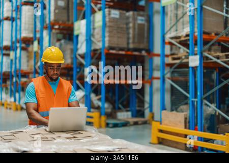 Schwarzer männlicher Arbeiter in einem Lager verwendet einen Laptop, um die Logistik zu verwalten. Das Bild zeigt moderne Technologie, Effizienz und sorgfältige Abläufe in einer Verteilungsumgebung. Geeignet für Funktionen für Unternehmenskommunikation, Lieferkette und digitale Lösungen. Stockfoto