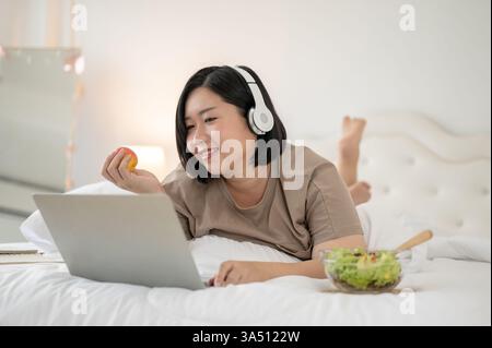 Glückliche asiatische Frau in Übergröße, die sich im Bett mit Kopfhörern und einem Laptop entspannt. Sie schaut sich einen Film an, surft online und plaudert aus einem gemütlichen Schlafzimmer. Perfekt für Lifestyle, Fernarbeit und Selbstpflege. Stockfoto