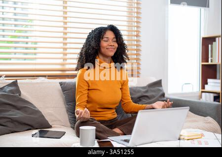 Eine ruhige schwarze Frau meditiert auf einem Sofa im Wohnzimmer und beginnt ihren Arbeitstag mit Achtsamkeit. Die friedliche Pose spiegelt Gleichgewicht und mentales Wohlbefinden in einer häuslichen Umgebung wider. Stockfoto