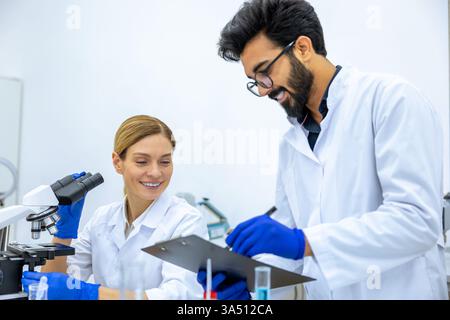 Fröhlicher indischer Wissenschaftler, der Labormantel und Latexhandschuhe trägt, macht Notizen, nachdem er einen Beispieltest in der Nähe von kaukasischen weiblichen Kollegen mit microsc gemacht hat... Stockfoto