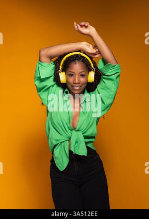 Glückliche Afroamerikanerin, die Kopfhörer trägt, tanzt und lächelt vor der Kamera. Dieses farbenfrohe Lifestyle-Image passt zu Musik-, Freizeit- und Wellness-Kampagnen mit einer lässigen, optimistischen Atmosphäre. Stockfoto