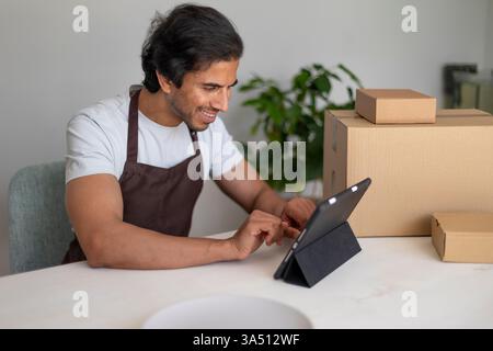 Ein indischer Mann in einem weißen Hemd und brauner Schürze benutzt eine Tablette in einer Heimpackstation. Umgeben von Boxen und Verpackungsmaterialien konzentriert er sich auf Online-Shopping-Logistik und kleine Geschäftsaufgaben in einem aufgeräumten Arbeitsbereich. Stockfoto
