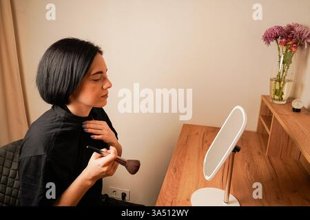 Die Frau konzentriert sich auf ihr Make-up und verwendet einen weichen Pinsel, um ihr Hautbild zu verbessern. Ihre zarten Züge spiegeln sich im Tischspiegel wider. Ihr dunkles Haar Stockfoto