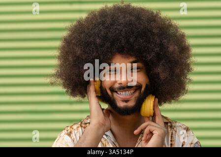 Bärtiger hispanischer Mann mit lockigem Haar schaut in die Kamera, während er kabellose Kopfhörer trägt. Dieses Studio-Porträt vermittelt eine fröhliche, stilvolle Atmosphäre, die für Lifestyle- und Musikinhalte geeignet ist. Stockfoto