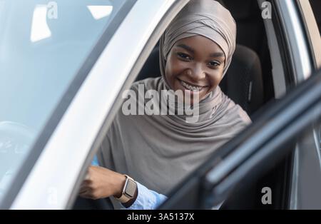 Die fröhliche schwarze muslimische Frau mit Hijab sitzt nach dem Kauf in ihrem neuen Auto und schaut in die Kamera. Dieses Lifestyle-Porträt fängt Besitz, Begeisterung und einen modernen Automobilmoment ein und ist ideal für Autohändler, Automobilmarketing und Lifestyle-Kampagnen. Stockfoto