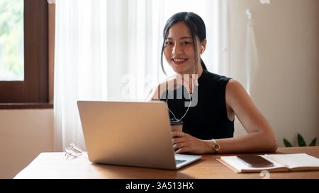 Asiatische Frau mit Ohrhörern arbeitet an ihrem Laptop an einem Tisch in einem Café. Ein Hinweis für Notebooks und drahtlose Verbindungen auf Remote-Arbeiten und Freiberufler. Stockfoto