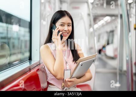 Eine positive asiatische Geschäftsfrau spricht am Telefon, während sie morgens in einem Skytrain pendelt. Die Szene des städtischen Nahverkehrs fängt einen modernen, hoffnungsvollen Start in den Tag ein. Geeignet für Reisen, Pendelfahrten und Konnektivität am Arbeitsplatz. Stockfoto