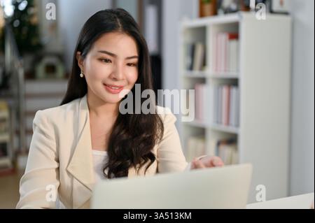 Lächelnde asiatische Geschäftsfrau sitzt an einem Schreibtisch in einem modernen Büro mit einem Laptop. Dieses professionelle Image unterstreicht weibliche Führungsqualitäten und Produktivität am Arbeitsplatz. Ideal für Themen zu Frauen in Unternehmen, Büros und thailändischer Arbeitskultur. Stockfoto
