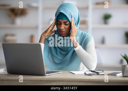 Kopfschmerzen-Konzept mit einer gestressten Schwarzen-muslimischen Freelancerin an ihrem Schreibtisch, die Migräneschmerzen erlebt. Die Szene vermittelt Stress, Gesundheit und Produktivität am Arbeitsplatz in einem modernen Büro. Geeignet für Geschäfts-, Wellness- und Diversitätsthemen. Stockfoto