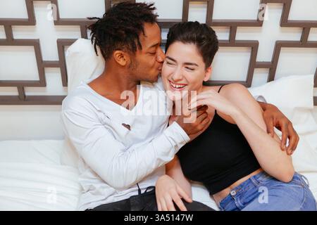 Eine liebevolle Szene eines afroamerikanischen Mannes, der seine kaukasische Freundin in einem gemütlichen Schlafzimmer umhüllt und küsst. Die Wärme und Zärtlichkeit des Paares betonen die romantische Intimität in einer häuslichen Umgebung. Ein vielseitiges Bild für Lifestyle, Beziehungen oder Geschichten am Morgen im Bett. Stockfoto