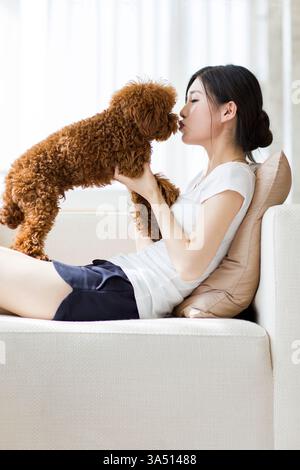 Eine junge Chinesin spielt zu Hause mit ihrem Pudel. Der gemütliche Moment im Haushalt vereint Lifestyle und Tierpflege in einem entspannten Wohnbereich. Perfekt für den Besitz von Haustieren, das Leben zu Hause und den modernen Lifestyle. Stockfoto