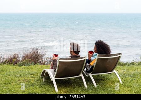 Schwarze Frau mit Afro-Frisur, die tagsüber auf einem Stuhl neben einem kaukasischen Freund in der Nähe des Meeres sitzt Stockfoto