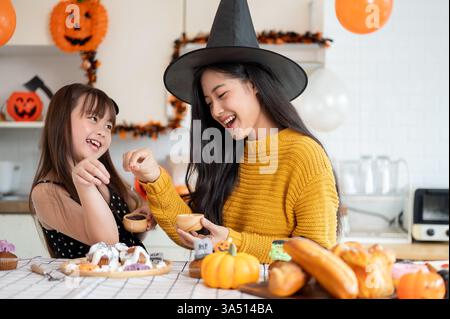 Ein glückliches asiatisches Mädchen und ihre Mutter in Halloween Kostümen lachen zusammen, während sie Halloween Cupcakes in der Küche dekorieren. Ideal für Feiertage, Familienfeiern und Backkampagnen. Stockfoto