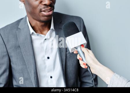 Zugeschnittenes Bild eines erfolgreichen schwarzen Geschäftsmannes, der während eines Interviews in ein Mikrofon spricht. Dieses professionelle Porträt eignet sich für Geschäfts-, Führungs-, Medien- und Kommunikationskontexte. Verwenden Sie diese Option für Artikel über öffentliche Vorträge, Pressekonferenzen oder Interviews mit Führungskräften. Stockfoto