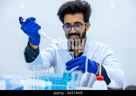 Ein lächelnder indischer Wissenschaftler in einem Labormantel und Handschuhen gießt chemische Flüssigkeit aus einer Pipette in einen Glaskolben an seinem Schreibtisch. Die Szene zeigt Laborarbeiten, Chemie und Biotechnologie, die für wissenschaftliche und pädagogische Funktionen geeignet sind. Es betont präzise Fokussierung und professionelle Arbeit in einer modernen Laborumgebung. Stockfoto