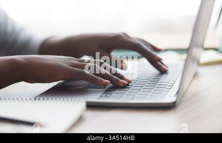 Nahaufnahme einer nicht erkennbaren Afro-Frau, die in einem modernen Büro auf einem Laptop tippt, präsentiert in einem Panoramablick. Vermittelt professionellen Fokus, digitale Arbeit und moderne Arbeitsumgebungen. Ideal für Business-Tech, Remote-Arbeiten und Images von Unternehmen. Stockfoto