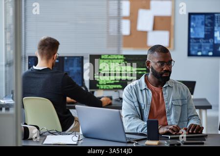 Porträt eines Afroamerikaners Programmiersoftware an einem Schreibtisch in einem modernen Büro. Die Komposition bietet einen klaren Kopierraum für Text, der sich für Technologie, Softwareentwicklung und IT-Themen eignet. Stockfoto