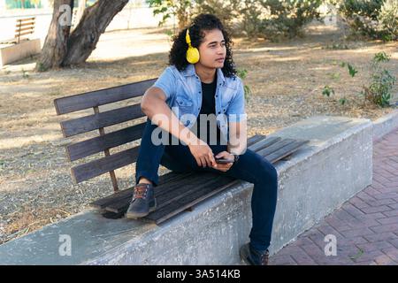 Ernsthafter hispanischer Mann mit Kopfhörern, der wegschaut und tagsüber mit einem Bein auf einer Holzbank im Park sitzt Stockfoto